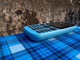 A blue feature phone with a numeric keypad rests on a surface covered with a blue plaid pattern. The background consists of a rough, textured wall with a mix of dark and light brown tones.