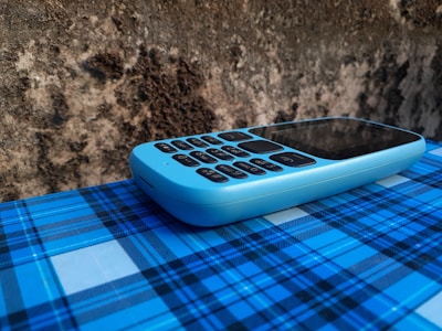 A blue feature phone with a numeric keypad rests on a surface covered with a blue plaid pattern. The background consists of a rough, textured wall with a mix of dark and light brown tones.