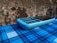 A blue feature phone with a numeric keypad rests on a surface covered with a blue plaid pattern. The background consists of a rough, textured wall with a mix of dark and light brown tones.