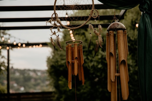 A close-up of a dreamcatcher and bamboo wind chimes hanging outdoors. The dreamcatcher is adorned with feathers and beads, while the bamboo chimes have a natural finish. Soft bokeh light strings and lush green foliage are visible in the background.