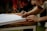 A person with red hair is signing a large, open book on a wooden table outdoors. The focus is on their hands and the pen, with the background slightly blurred.