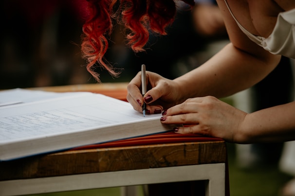 A person with red hair is signing a large, open book on a wooden table outdoors. The focus is on their hands and the pen, with the background slightly blurred.