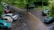 A flooded street scene with parked cars and a person holding an umbrella walking along the watery road. Surrounding trees are lush and green, and the ground is wet from rain. Several vehicles are parked along the street, with water covering parts of the road.