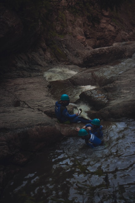 A group of three people wearing blue helmets and wetsuits are positioned near a small waterfall in a rocky, forested area. Two of them seem to be in the water while the third one sits on a rock.
