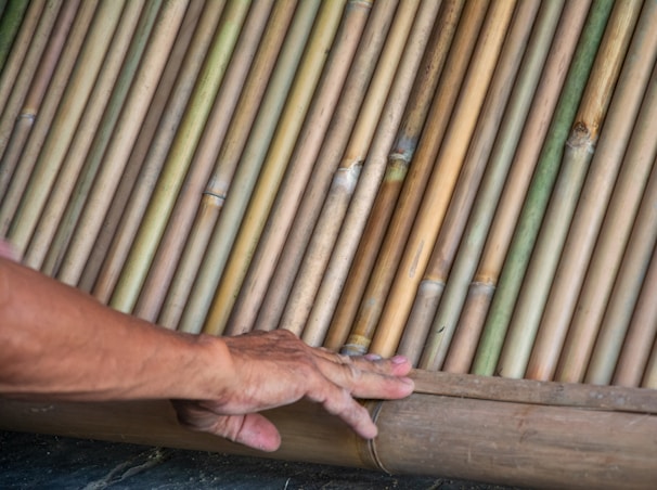 A hand rests gently on a surface made of closely arranged bamboo sticks. The bamboo sticks vary in color from light green to golden brown, showcasing natural textures and variations.