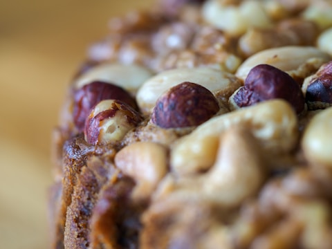 A close-up view of a baked item topped with an assortment of nuts, including cashews, hazelnuts, and possibly almonds. The surface has a rich texture, and the nuts are slightly toasted, giving a rustic and hearty appearance.