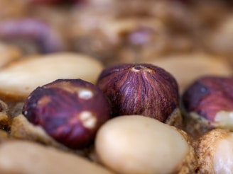 Close-up view of various nuts, including hazelnuts with a brown shell and peeled almonds with a light beige color. The texture of the nuts is detailed and prominent.