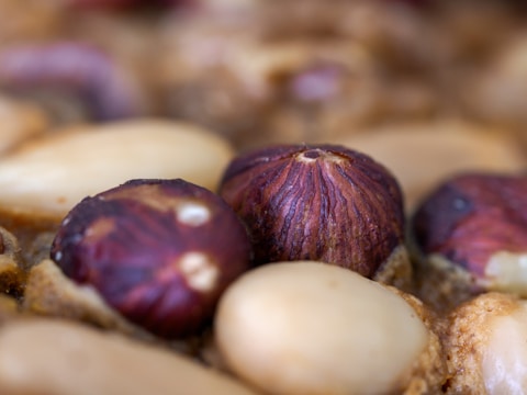 Close-up view of various nuts, including hazelnuts with a brown shell and peeled almonds with a light beige color. The texture of the nuts is detailed and prominent.