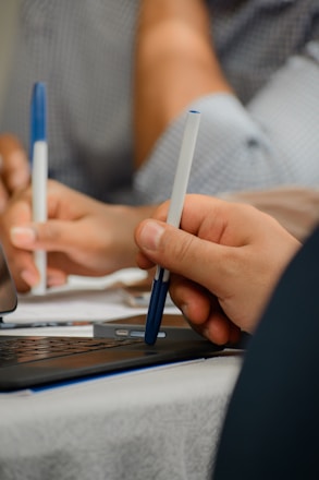 A close-up of hands editing a printed registration list with a pen and laptop in the background.