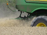 A green agricultural machine is in the midst of harvesting a field of wheat or a similar crop. Dust and chaff fill the air as the machine moves through the field, kicking up wheat stalks and cutting through the plants. The large wheel is prominently visible, with its brand name readable on the side.