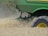 A green agricultural machine is in the midst of harvesting a field of wheat or a similar crop. Dust and chaff fill the air as the machine moves through the field, kicking up wheat stalks and cutting through the plants. The large wheel is prominently visible, with its brand name readable on the side.