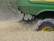 A green agricultural machine is in the midst of harvesting a field of wheat or a similar crop. Dust and chaff fill the air as the machine moves through the field, kicking up wheat stalks and cutting through the plants. The large wheel is prominently visible, with its brand name readable on the side.