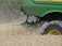 A green agricultural machine is in the midst of harvesting a field of wheat or a similar crop. Dust and chaff fill the air as the machine moves through the field, kicking up wheat stalks and cutting through the plants. The large wheel is prominently visible, with its brand name readable on the side.