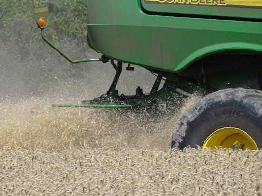 A green agricultural machine is in the midst of harvesting a field of wheat or a similar crop. Dust and chaff fill the air as the machine moves through the field, kicking up wheat stalks and cutting through the plants. The large wheel is prominently visible, with its brand name readable on the side.