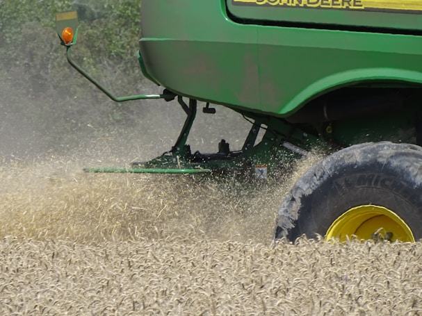 A green agricultural machine is in the midst of harvesting a field of wheat or a similar crop. Dust and chaff fill the air as the machine moves through the field, kicking up wheat stalks and cutting through the plants. The large wheel is prominently visible, with its brand name readable on the side.