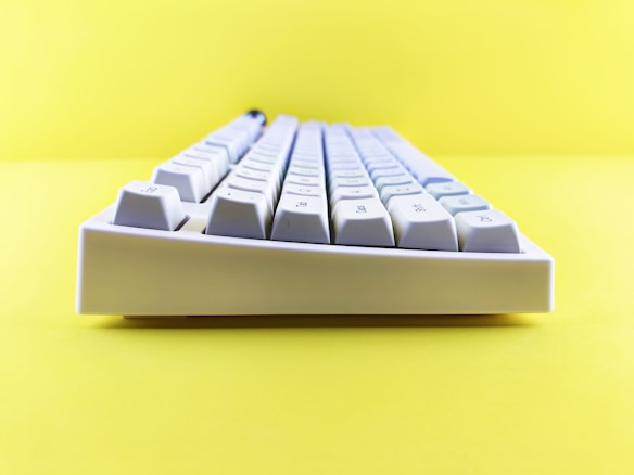 A white mechanical keyboard is positioned against a bright yellow background, creating a striking contrast. The keyboard is viewed from a side angle, highlighting the profile of the keys.