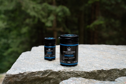 Two jars labeled 'Crystalweed Amnesia Canna' are placed on a large, flat stone surface, with a forested background. They appear to be products related to CBD or cannabis.
