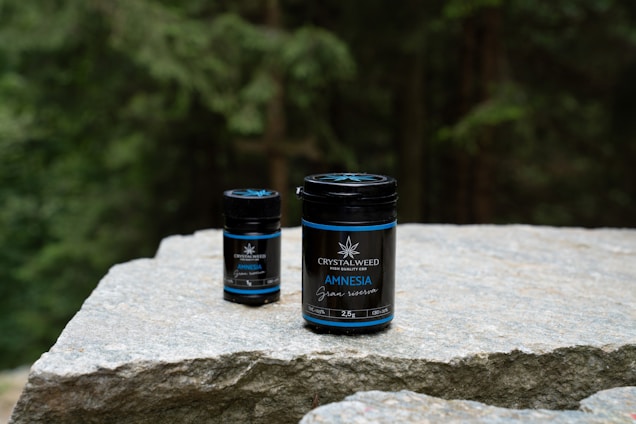 Two jars labeled 'Crystalweed Amnesia Canna' are placed on a large, flat stone surface, with a forested background. They appear to be products related to CBD or cannabis.