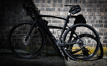 A sleek black road bike leans against a weathered brick wall, casting a sharp shadow on the rough surface. The bike's modern design features aerodynamic lines and a high-performance frame, ideal for speed and agility.
