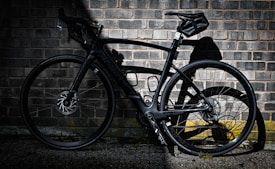 A sleek black road bike leans against a weathered brick wall, casting a sharp shadow on the rough surface. The bike's modern design features aerodynamic lines and a high-performance frame, ideal for speed and agility.