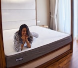 A person is lying on a large mattress in a cozy bedroom setting, resting their chin on their hands with a relaxed expression. The room features a wooden bed frame, soft white and gray bedding, a bedside table, and tall, elegant curtains that let in natural light.