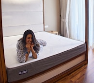 A person is lying on a large mattress in a cozy bedroom setting, resting their chin on their hands with a relaxed expression. The room features a wooden bed frame, soft white and gray bedding, a bedside table, and tall, elegant curtains that let in natural light.