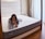 A person is lying on a large mattress in a cozy bedroom setting, resting their chin on their hands with a relaxed expression. The room features a wooden bed frame, soft white and gray bedding, a bedside table, and tall, elegant curtains that let in natural light.