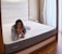 A person is lying on a large mattress in a cozy bedroom setting, resting their chin on their hands with a relaxed expression. The room features a wooden bed frame, soft white and gray bedding, a bedside table, and tall, elegant curtains that let in natural light.