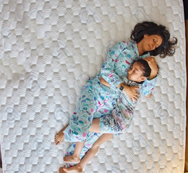 A woman and a child are lying on a mattress, both wearing pajamas. The woman is holding the child, who is resting peacefully in her arms. The mattress has a textured pattern, and they are surrounded by a sense of calm and comfort.