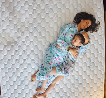 A woman and a child are lying on a mattress, both wearing pajamas. The woman is holding the child, who is resting peacefully in her arms. The mattress has a textured pattern, and they are surrounded by a sense of calm and comfort.