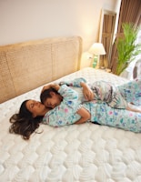 A woman and a child are lying on a bed, embracing each other. They are wearing pajamas with colorful patterns. The room is well-lit with a lamp on the bedside table next to a plant. The bed has a woven headboard and a white quilted mattress. There is a large mirror near the window with curtains drawn aside.