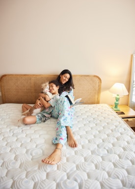 A woman and a child are sitting on a large bed with a rattan headboard, appearing joyful and relaxed. The child is hugging a small dog, and there is an open book in the woman's hand. A lamp on a bedside table provides a warm light in the otherwise neutral-colored room.