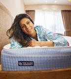 A woman wearing patterned pajamas is lying on a blue and white mattress with a wooden bed frame. She appears to be smiling and resting her arms on the mattress. Behind her, there are brown curtains and a window allowing soft natural light to illuminate the room.