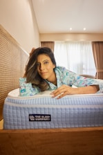 A person is lying on a bed, leaning on the mattress with a relaxed expression. The individual is wearing patterned pajamas. The room has a modern style with a wooden headboard and curtains partially open to reveal a window. The mattress is branded 'The Sleep Company' and has a textured blue and white top.