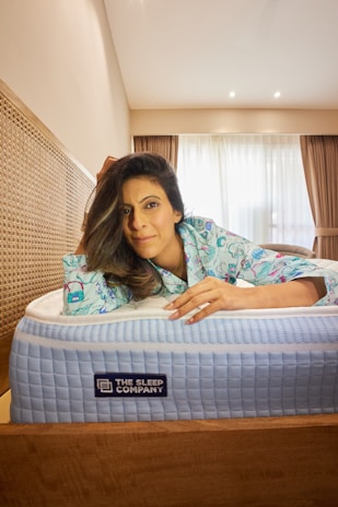 A person is lying on a bed, leaning on the mattress with a relaxed expression. The individual is wearing patterned pajamas. The room has a modern style with a wooden headboard and curtains partially open to reveal a window. The mattress is branded 'The Sleep Company' and has a textured blue and white top.