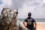 A person stands on a beach wearing a blue shirt with the words 'My Generation Will Save The Planet.' In front of them is a large art installation resembling a frog, constructed from netting and filled with plastic waste. The sea and a cloudy sky serve as a backdrop, highlighting the environmental message.