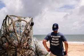 A person stands on a beach wearing a blue shirt with the words 'My Generation Will Save The Planet.' In front of them is a large art installation resembling a frog, constructed from netting and filled with plastic waste. The sea and a cloudy sky serve as a backdrop, highlighting the environmental message.