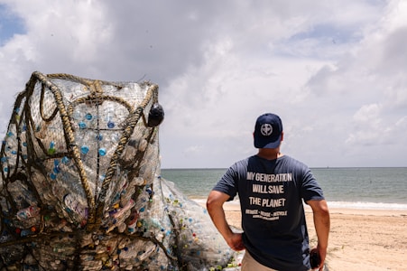 A person stands on a beach wearing a blue shirt with the words 'My Generation Will Save The Planet.' In front of them is a large art installation resembling a frog, constructed from netting and filled with plastic waste. The sea and a cloudy sky serve as a backdrop, highlighting the environmental message.