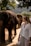A woman stands on a dirt path next to a group of elephants in a natural, wooded environment. The scene is filled with green foliage and trees, and the woman appears to be observing the elephants closely. She is holding a folded cloth and a phone, dressed casually in light-colored clothing. The lighting suggests a sunny day, with the background featuring mountains covered in greenery.