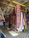 An array of colorful textiles and hammocks displayed in a market setting. The textiles feature vibrant floral and geometric patterns, with a focus on bold colors like pink, blue, and orange. Several hammocks are hung alongside the textiles, and the display is under a shelter made of wooden beams and a corrugated metal roof.
