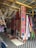 An array of colorful textiles and hammocks displayed in a market setting. The textiles feature vibrant floral and geometric patterns, with a focus on bold colors like pink, blue, and orange. Several hammocks are hung alongside the textiles, and the display is under a shelter made of wooden beams and a corrugated metal roof.