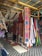 An array of colorful textiles and hammocks displayed in a market setting. The textiles feature vibrant floral and geometric patterns, with a focus on bold colors like pink, blue, and orange. Several hammocks are hung alongside the textiles, and the display is under a shelter made of wooden beams and a corrugated metal roof.