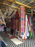 An array of colorful textiles and hammocks displayed in a market setting. The textiles feature vibrant floral and geometric patterns, with a focus on bold colors like pink, blue, and orange. Several hammocks are hung alongside the textiles, and the display is under a shelter made of wooden beams and a corrugated metal roof.