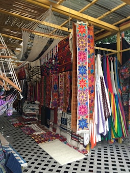 An array of colorful textiles and hammocks displayed in a market setting. The textiles feature vibrant floral and geometric patterns, with a focus on bold colors like pink, blue, and orange. Several hammocks are hung alongside the textiles, and the display is under a shelter made of wooden beams and a corrugated metal roof.