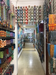 A colorful shop aisle filled with various traditional beaded items hanging on both side walls. The beads are arranged in intricate patterns and vibrant colors, creating an artistic and cultural ambiance. The overhead lighting casts a warm glow on the items, enhancing their vivid hues.