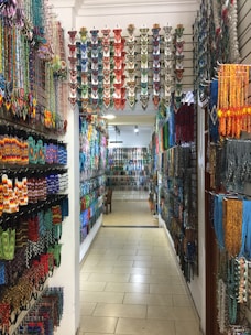 A colorful shop aisle filled with various traditional beaded items hanging on both side walls. The beads are arranged in intricate patterns and vibrant colors, creating an artistic and cultural ambiance. The overhead lighting casts a warm glow on the items, enhancing their vivid hues.