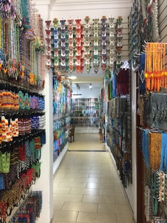 A colorful shop aisle filled with various traditional beaded items hanging on both side walls. The beads are arranged in intricate patterns and vibrant colors, creating an artistic and cultural ambiance. The overhead lighting casts a warm glow on the items, enhancing their vivid hues.