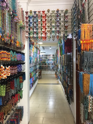 A colorful shop aisle filled with various traditional beaded items hanging on both side walls. The beads are arranged in intricate patterns and vibrant colors, creating an artistic and cultural ambiance. The overhead lighting casts a warm glow on the items, enhancing their vivid hues.