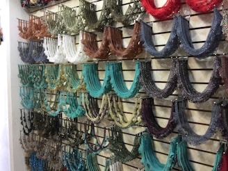 An assortment of colorful beaded necklaces is neatly displayed on a slatted wall. The necklaces are arranged by color groups, including shades of brown, white, black, teal, and red, creating a visually appealing pattern.