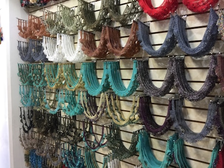 An assortment of colorful beaded necklaces is neatly displayed on a slatted wall. The necklaces are arranged by color groups, including shades of brown, white, black, teal, and red, creating a visually appealing pattern.
